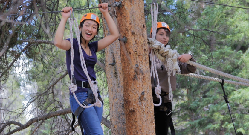 Students wearing safety gear is secured by ropes as they smile at the camera during a ropes course. 